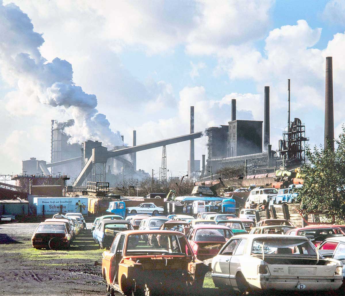 Was verbirgt sich hinter Begriffen wie „Löschen“ und „Wolkenmachen“ auf einer Kokerei? Und was überhaupt passiert in einer Kokerei? In einer Kokerei wird aus Kohle mittels eines trockenen Destillationsverfahrens Koks und Rohgas erzeugt. Es werden die flüchtigen Bestandteile in der Kohle durch das Erhitzen auf eine Temperatur von bis zu 1400 Grad pyrolysiert, freigesetzt und abgesaugt. Nach diesem Prozess wird der Koks aus der Batterie ins Freie gedrückt. Bei den dann noch herrschenden Temperaturen würde er beim Kontakt mit dem Luftsauerstoff verbrennen. Um das zu verhindern, muss er schnell gelöscht und abgekühlt werden. Dabei entsteht Wasserdampf, die Wolke. Genau diesen Moment fing Manfred Ehrich 1985 an der Kokerei Osterfeld in Oberhausen ein, mit Capri II, Manta B und Co. als stummen Zeugen auf dem angrenzenden Schrottplatz. Was verbirgt sich hinter Begriffen wie „Löschen“ und „Wolkenmachen“ auf einer Kokerei? Und was überhaupt passiert in einer Kokerei? In einer Kokerei wird aus Kohle mittels eines trockenen Destillationsverfahrens Koks und Rohgas erzeugt. Es werden die flüchtigen Bestandteile in der Kohle durch das Erhitzen auf eine Temperatur von bis zu 1400 Grad pyrolysiert, freigesetzt und abgesaugt. Nach diesem Prozess wird der Koks aus der Batterie ins Freie gedrückt. Bei den dann noch herrschenden Temperaturen würde er beim Kontakt mit dem Luftsauerstoff verbrennen. Um das zu verhindern, muss er schnell gelöscht und abgekühlt werden. Dabei entsteht Wasserdampf, die Wolke. Genau diesen Moment fing Manfred Ehrich 1985 an der Kokerei Osterfeld in Oberhausen ein, mit Capri II, Manta B und Co. als stummen Zeugen auf dem angrenzenden Schrottplatz.